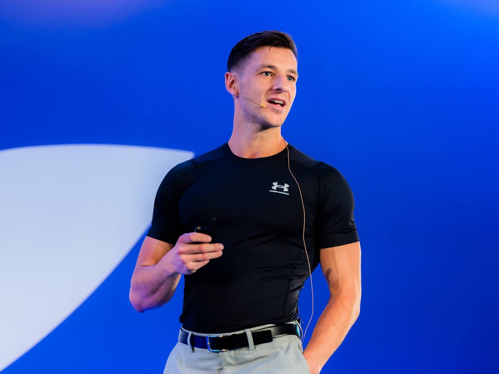 Delano speaking on stage against a blue backdrop, headset microphone