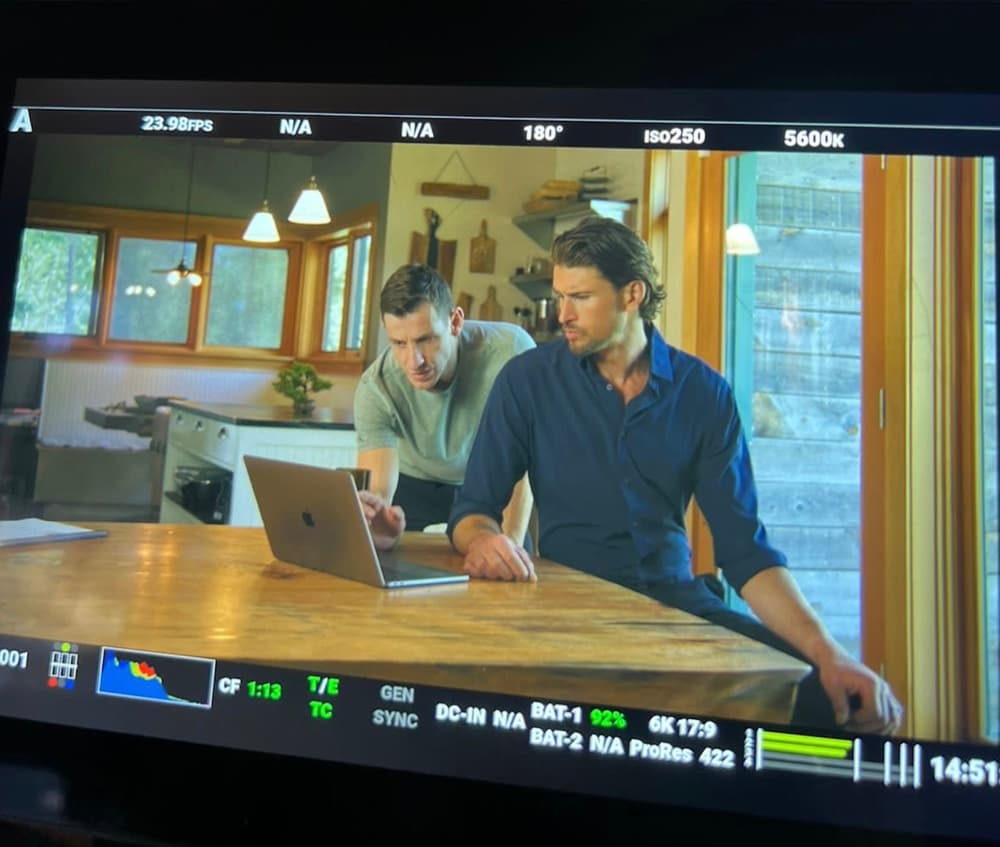 Camera-monitor view of Delano directing a kitchen scene, reviewing a laptop with an actor